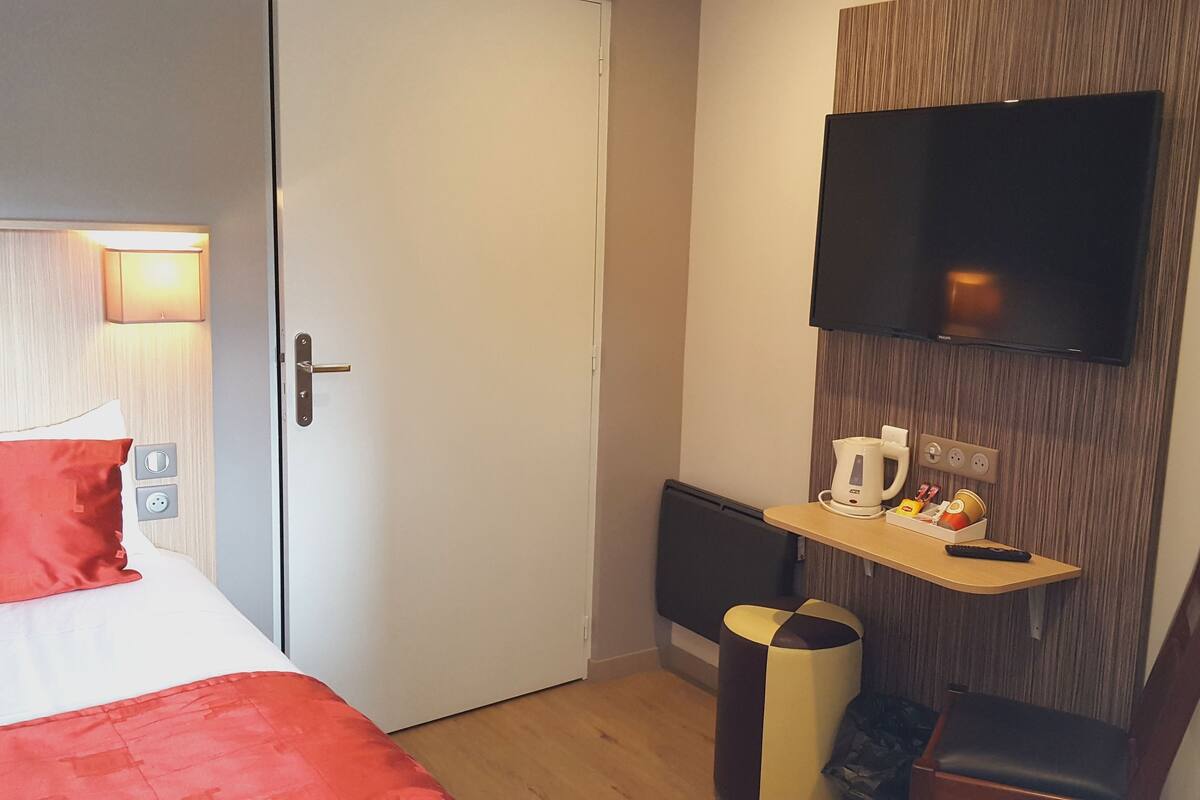 Habitación de hotel con una cama, un televisor en la pared, una mesita con una tetera y una bandeja, y una silla.