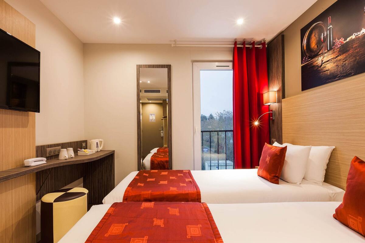 Habitación de hotel con dos camas, televisión, escritorio con teléfono y tetera, y balcón con cortinas rojas.
