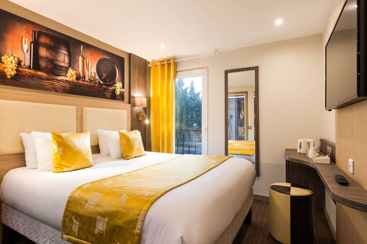 Habitación de hotel con una cama grande, un cuadro sobre la cama, un espejo, un escritorio con una computadora y una ventana con cortinas.
