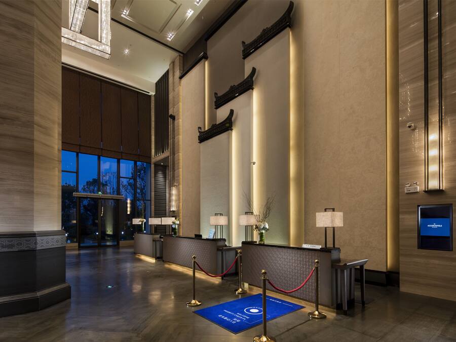 Un vestíbulo de hotel con mostrador de recepción, una alfombra azul con un logotipo y un ventanal con vistas a árboles.