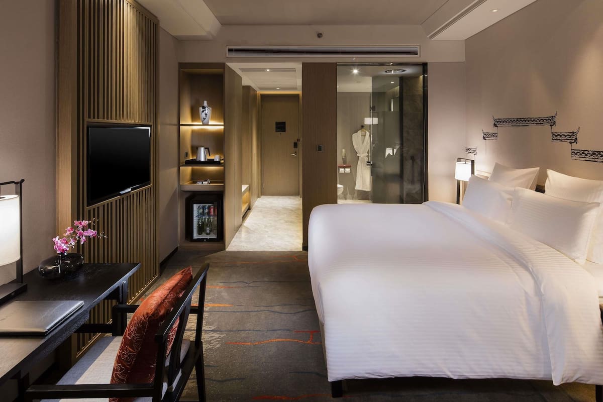 Une chambre dâhĂŽtel Ă©quipĂ©e dâun lit, dâun bureau avec un ordinateur portable, dâune tĂ©lĂ©vision, dâun mini-bar et dâune salle de bain visible Ă travers une porte en verre.
