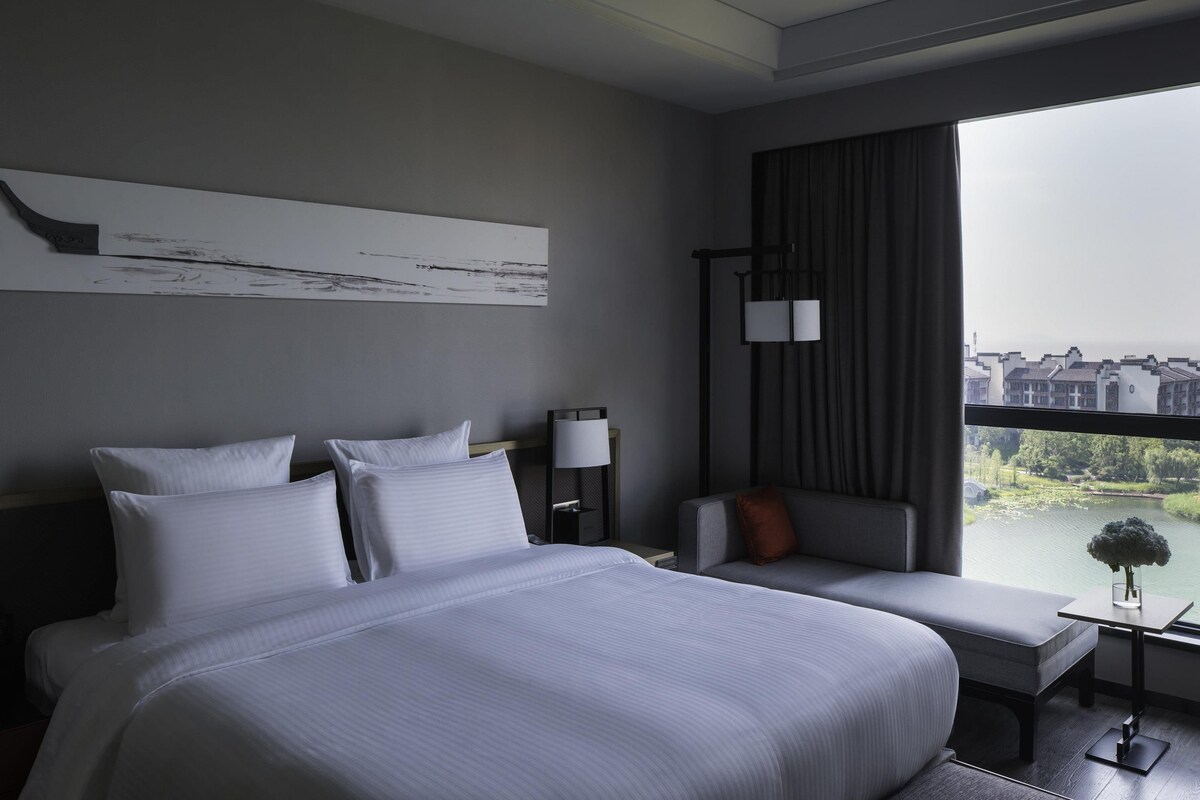 A modern hotel room with a large bed, a seating area, and a view of buildings through a window.