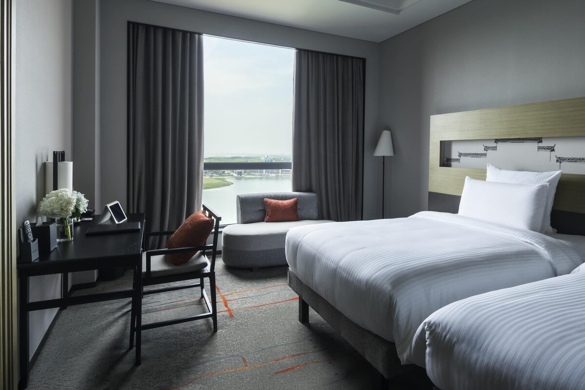 A hotel room with a bed, a desk, a chair, a lamp, and a view of a body of water.