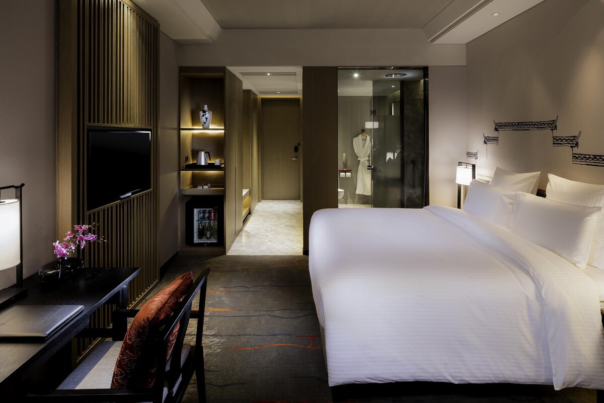 A hotel room with a bed, a desk, a TV, a minibar, and a bathroom visible through a glass door.