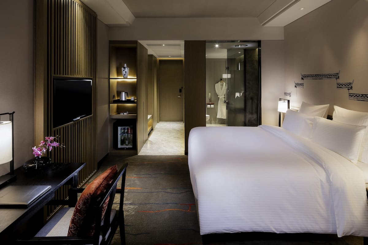 A hotel room with a bed, a desk, a TV, a minibar, and a bathroom visible through a glass door.