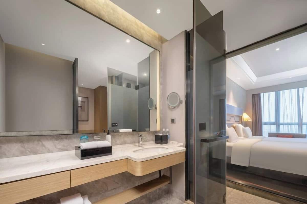 A modern hotel bathroom with a large mirror, two sinks, a glass shower enclosure, and a bed visible through the shower door.