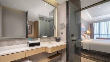 Un baño de hotel moderno con un espejo grande, dos lavamanos, una cabina de ducha de vidrio y una cama visible a través de la puerta de la ducha.