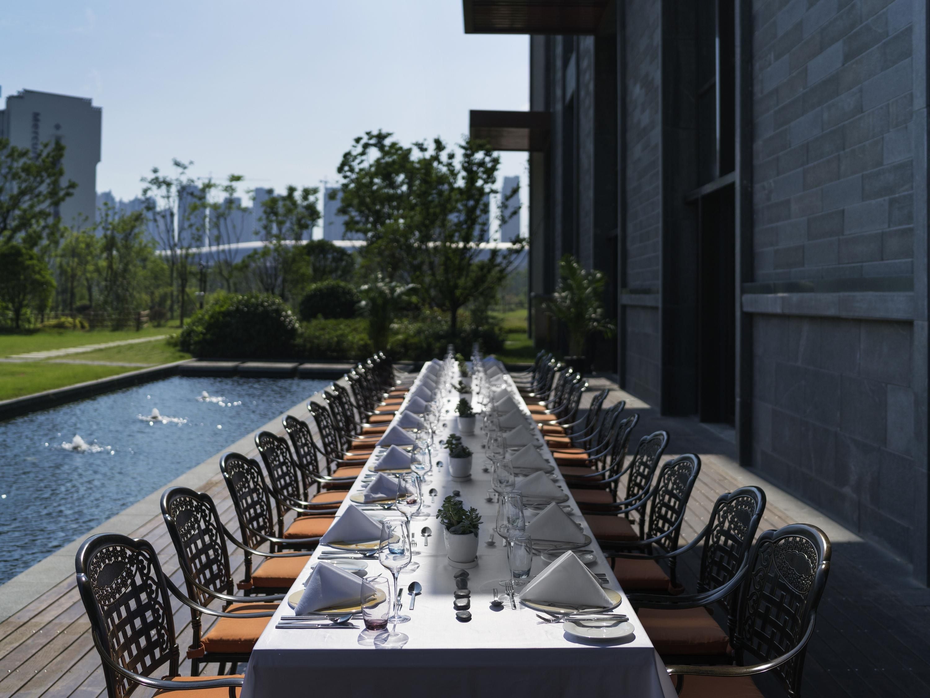 A long dining table set for a meal with chairs on both sides, overlooking a water feature and a cityscape.