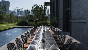 A long dining table set for a meal with chairs on both sides, overlooking a water feature and a cityscape.
