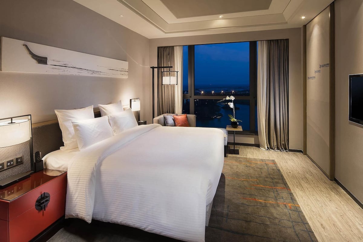 A hotel room with a large bed, a desk with a lamp, a TV mounted on the wall, and a view of the city at night.