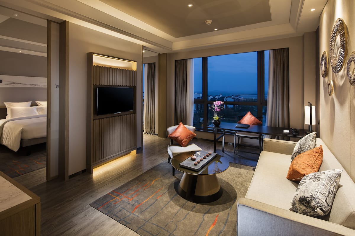 A hotel room with a bed, a TV, a sofa, a dining table, and a view of the city.