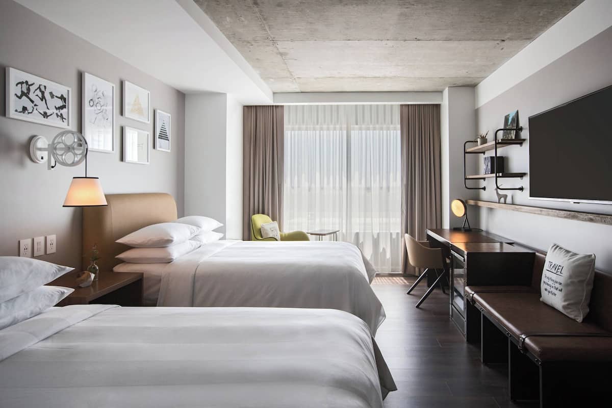 A hotel room with two beds, a desk, a chair, a TV, and artwork on the walls.