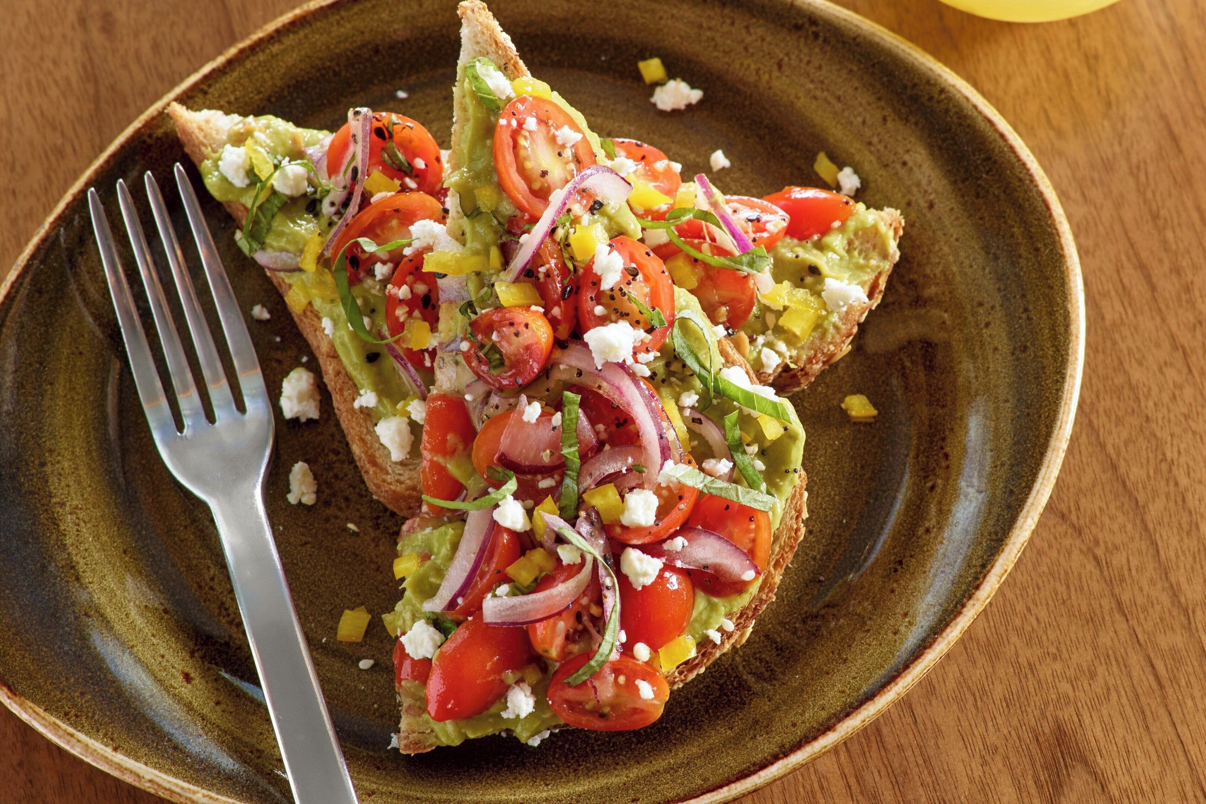 Ein Teller mit Toast, belegt mit Avocado, Tomaten, Zwiebeln und Feta-Käse.