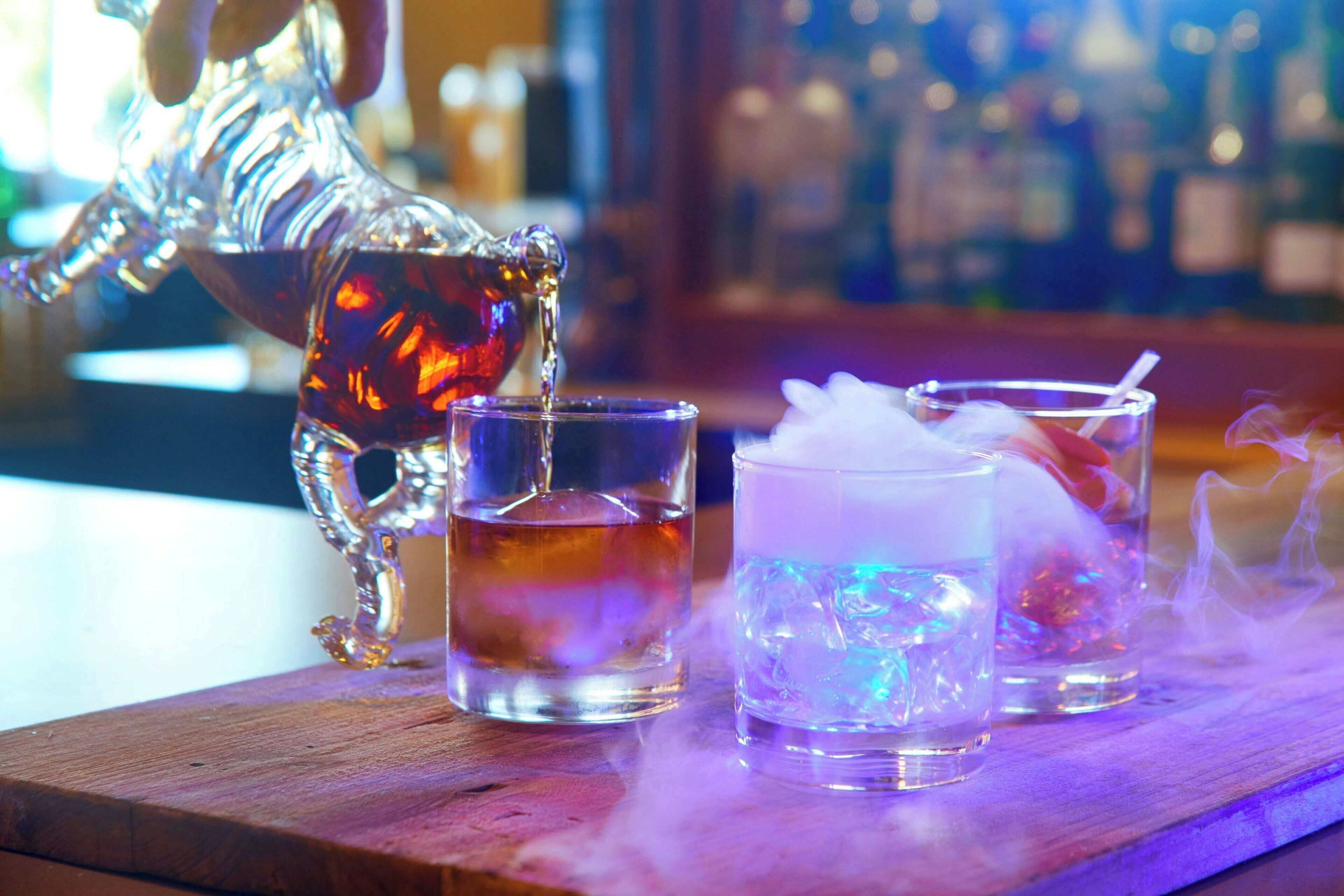 Une scène de bar avec un pichet décoratif servant une boisson dans un verre, accompagné d’un verre à whisky et d’un grand verre à cocktail rempli de glace et muni d’une paille.