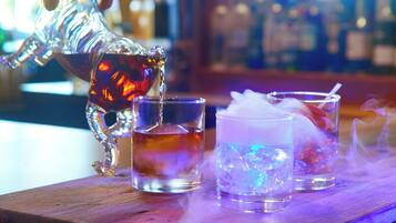 Un bar con una jarra decorativa sirviendo una bebida en un vaso, junto a un vaso bajo y un vaso alto con hielo y una pajita.