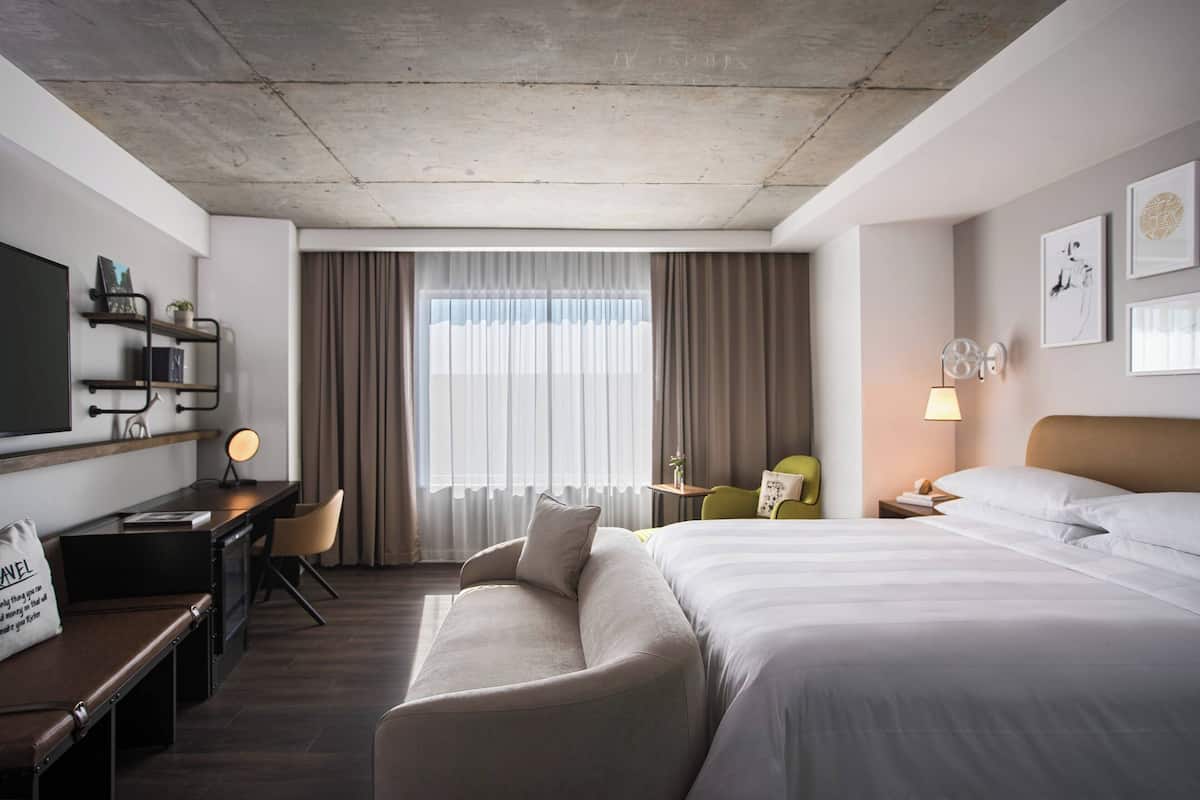 A modern hotel room with a large bed, a desk with a chair, a sofa, and a small table with a lamp.