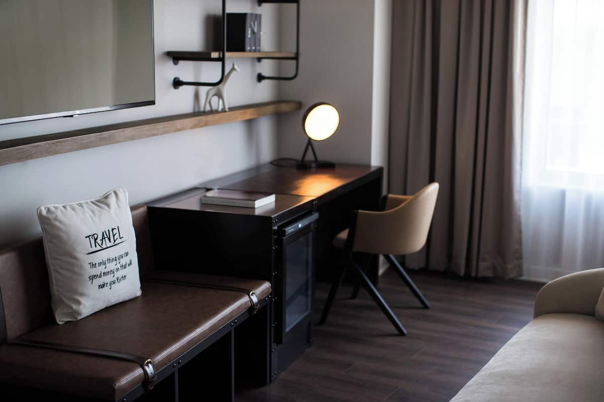 A hotel room with a wooden desk, a chair, a cushioned bench, a bedside table, a lamp, and a mirror on the wall.