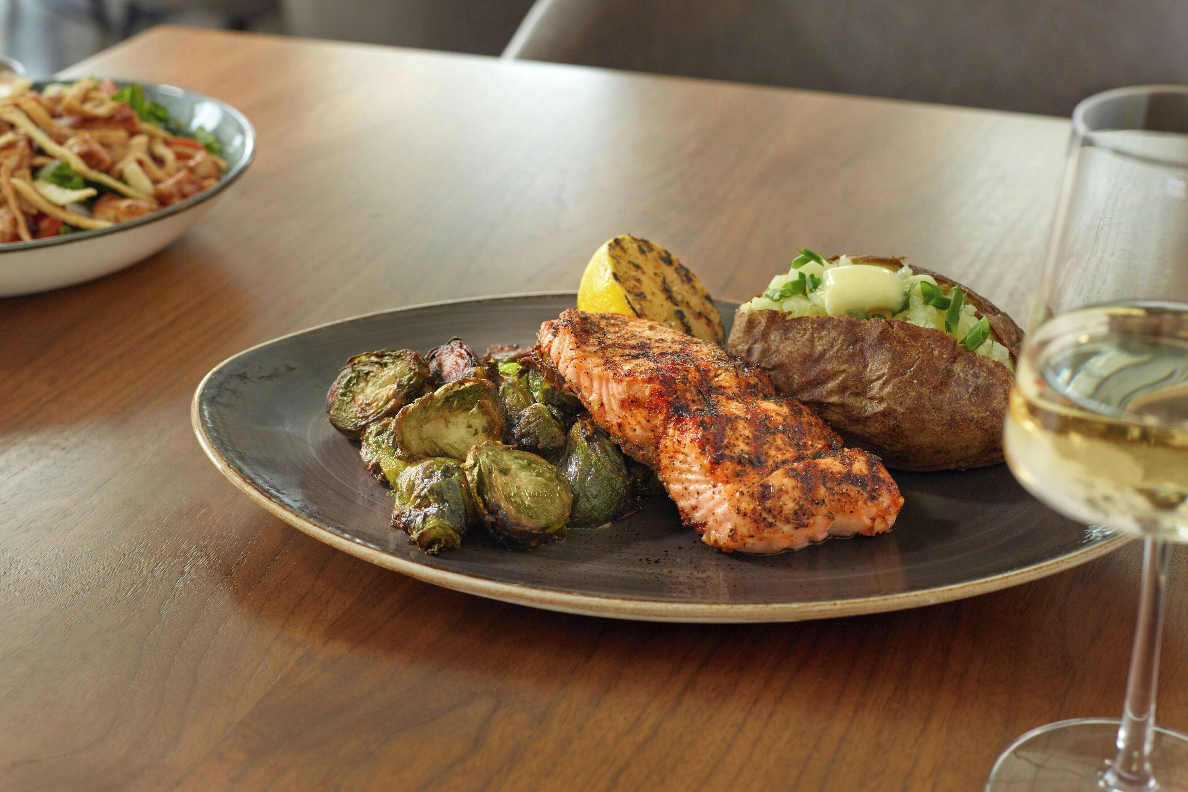 Un plato con salmón a la parrilla, brócoli belga asado y una patata al horno, acompañado de una copa de vino blanco sobre una mesa de madera.
