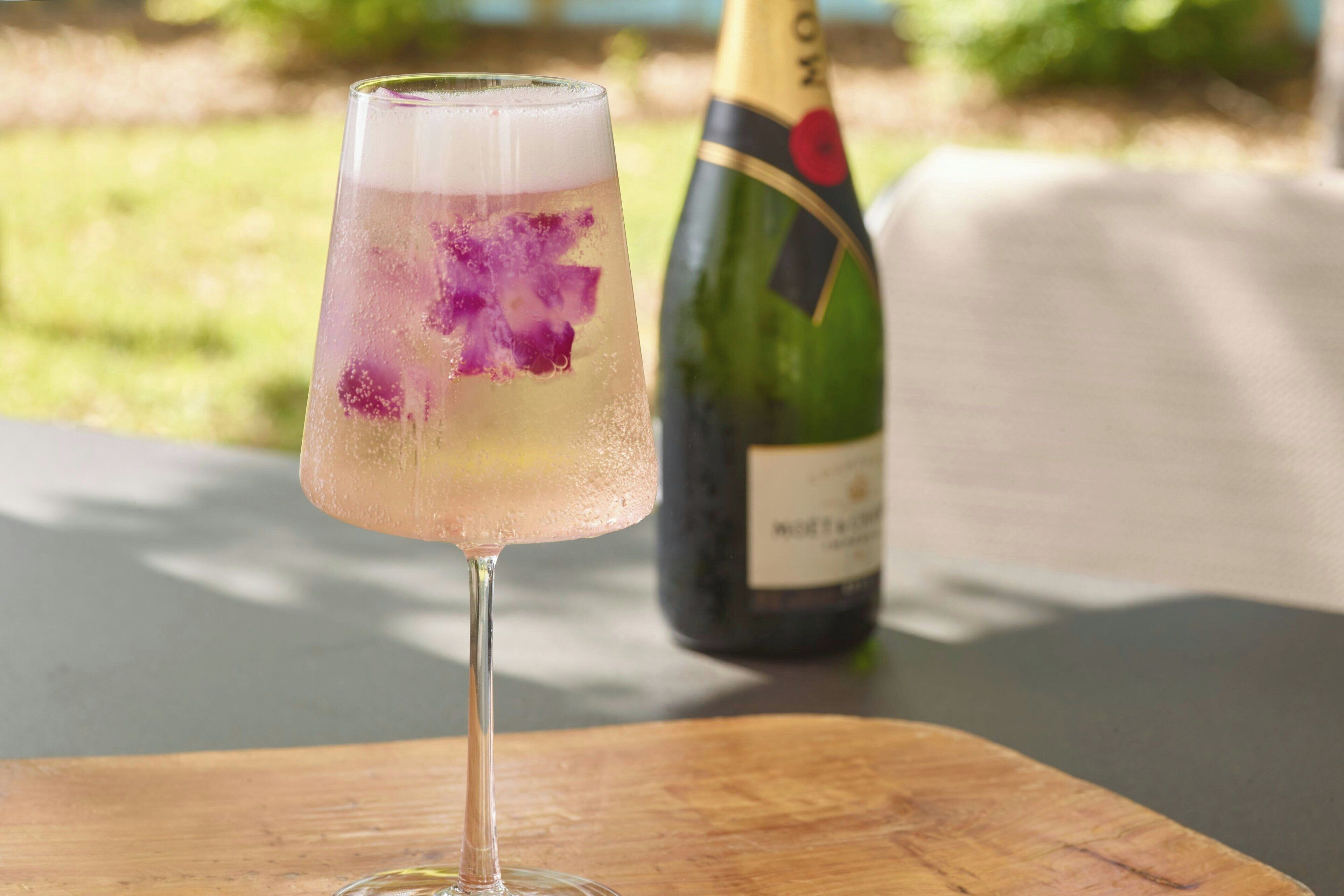 A glass of cocktail with a floral garnish on a wooden table, with a bottle of champagne in the background.