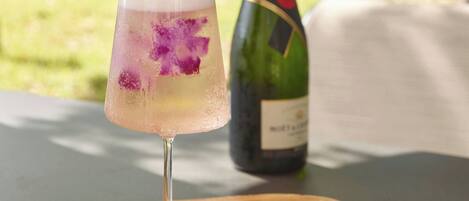 A glass of cocktail with a floral garnish on a wooden table, with a bottle of champagne in the background.