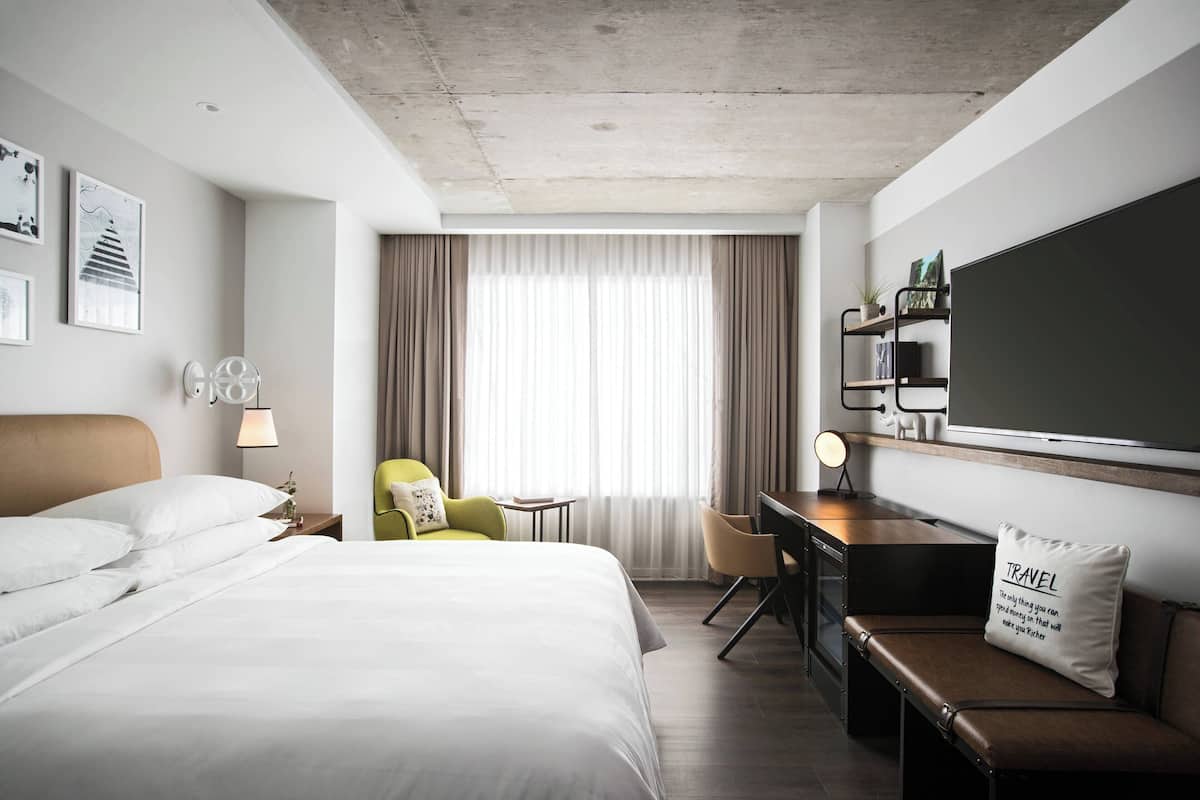A hotel room with a large bed, a desk, a chair, a small table, and a bookshelf.
