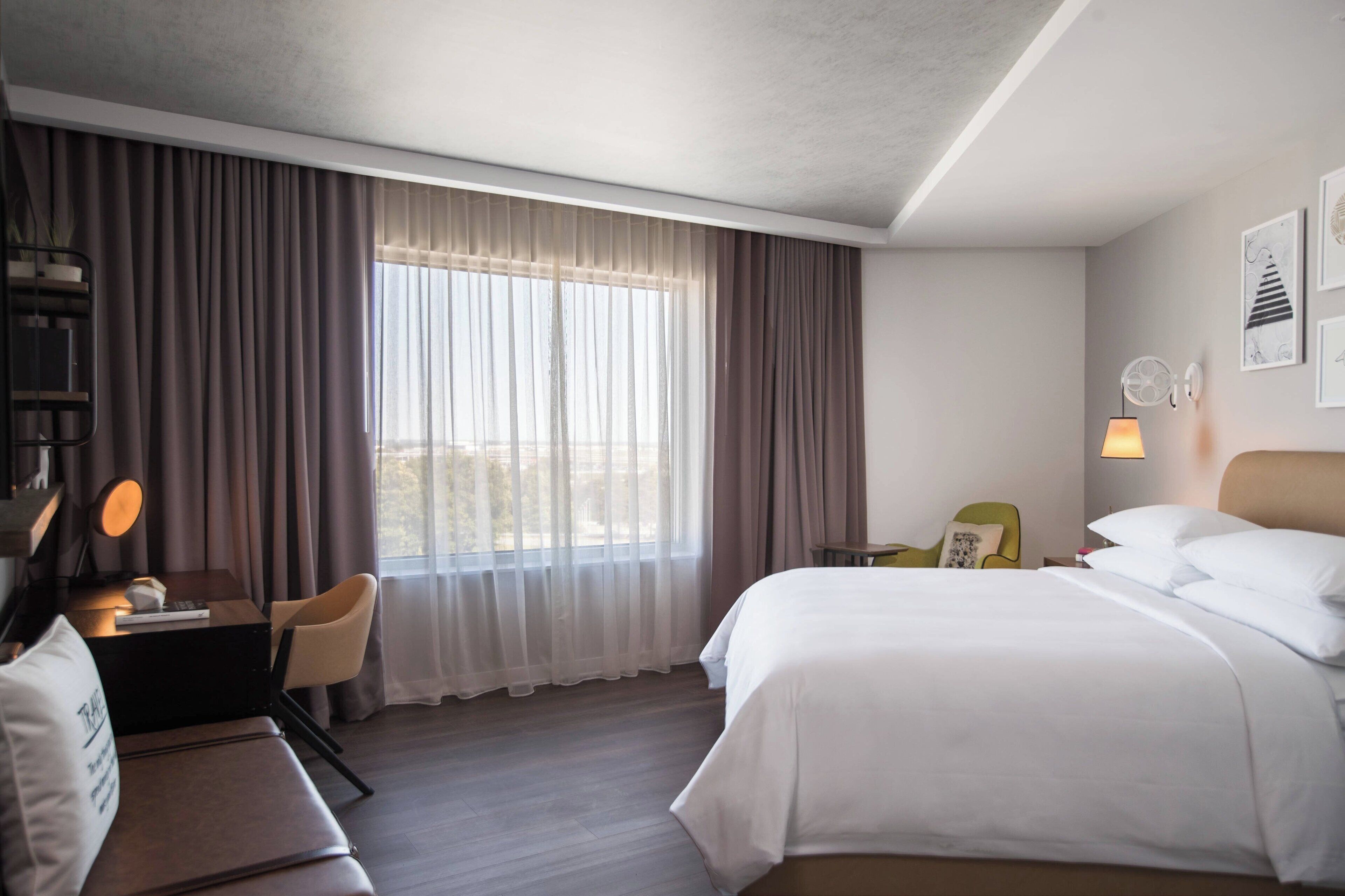 Une chambre d’hôtel avec un grand lit, un bureau avec une lampe, une chaise et une vue sur l’extérieur à travers des rideaux légers.