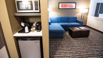 Habitación de hotel compacta con kitchenette, microondas, cafetera, refrigerador pequeño, sofá azul, pequeño reposapiés y un cuadro en la pared.