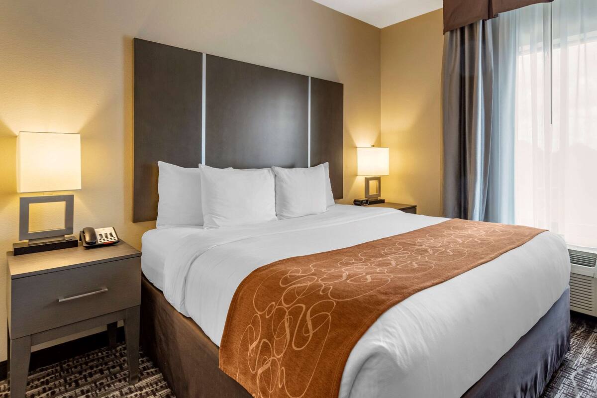 Une chambre d’hôtel avec un grand lit, deux tables de chevet avec des lampes, une petite table avec un téléphone et une fenêtre avec des rideaux.
