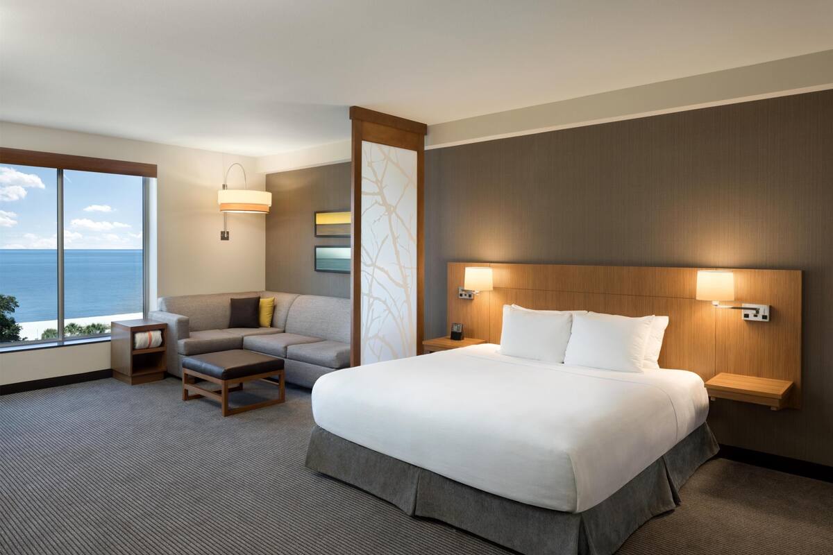 Quarto de hotel com uma cama grande, um sofá, uma mesa pequena e vista para o oceano.