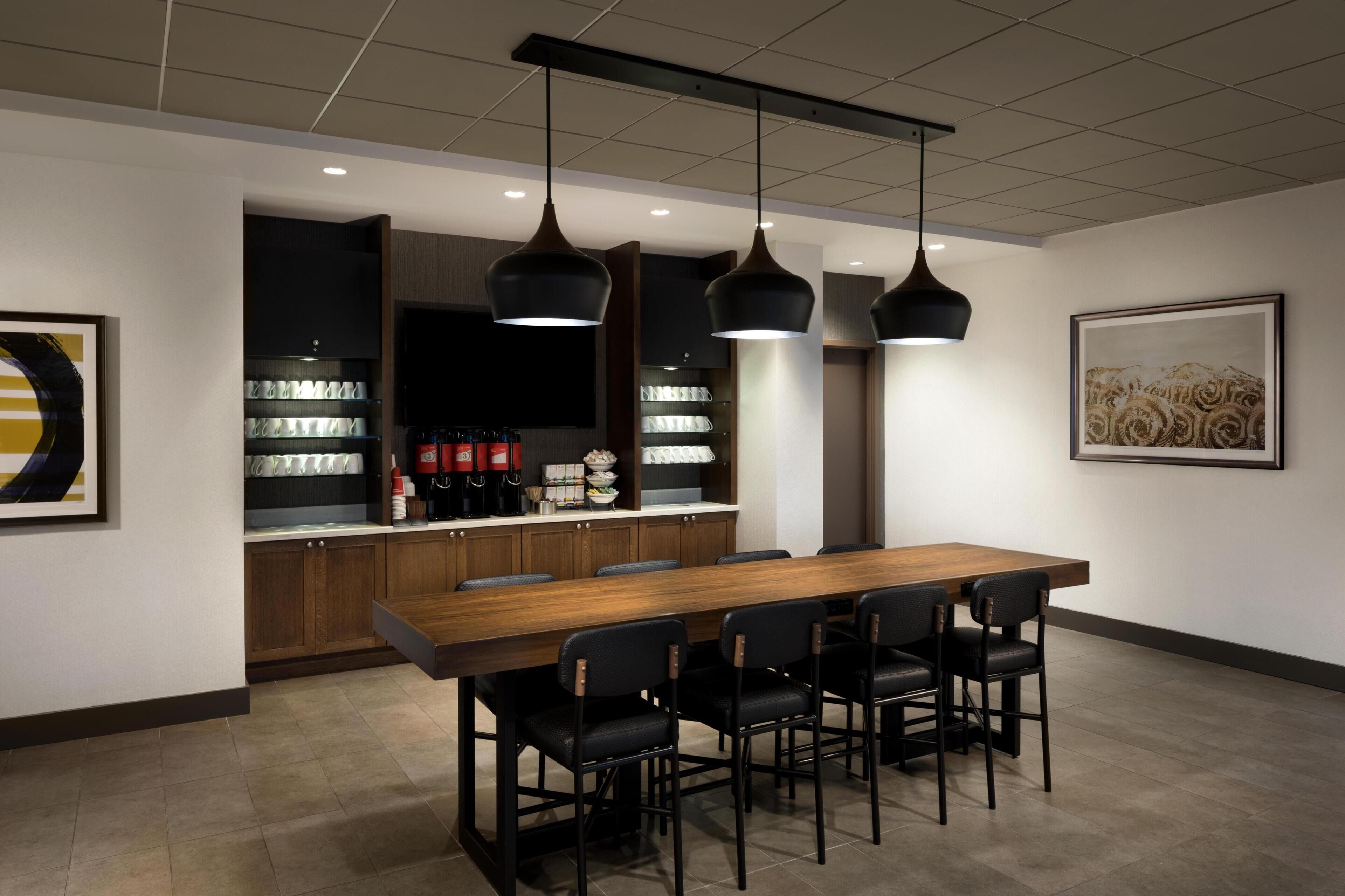A modern dining area with a long wooden table, black chairs, pendant lights, and a kitchen area with shelves.
