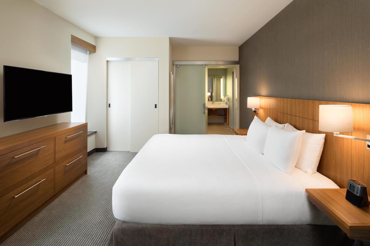 Quarto de hotel com uma cama grande, televisão de tela plana, cômoda de madeira e mesa de cabeceira com abajur.