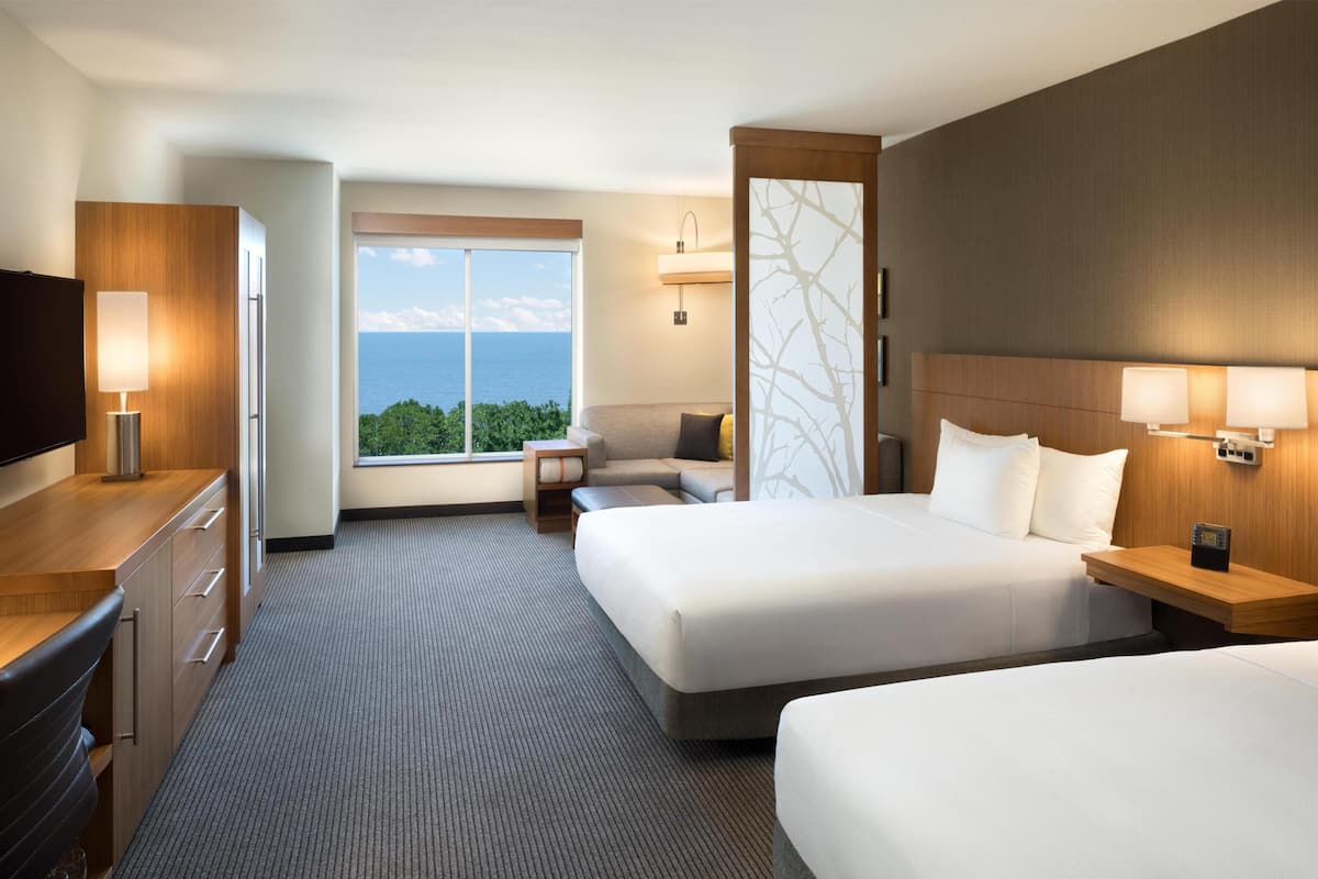 Quarto de hotel com duas camas, uma escrivaninha com televisão, uma janela com vista para o oceano e um abajur fixado na parede.