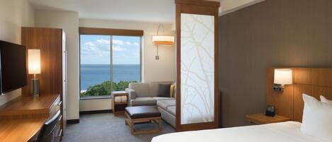 A hotel room with a bed, a desk, a TV, a sofa, and a view of the ocean.