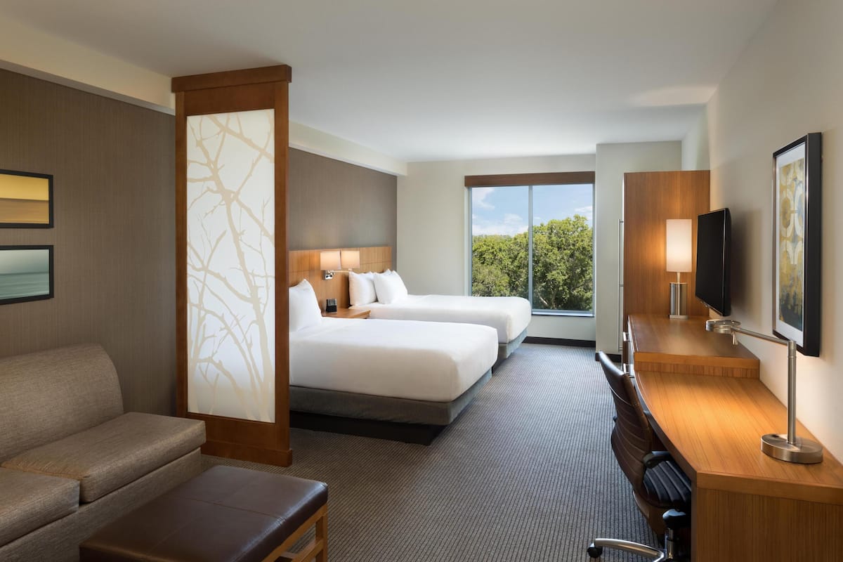 Quarto de hotel com duas camas, um sofá, uma escrivaninha, uma televisão e uma janela grande.