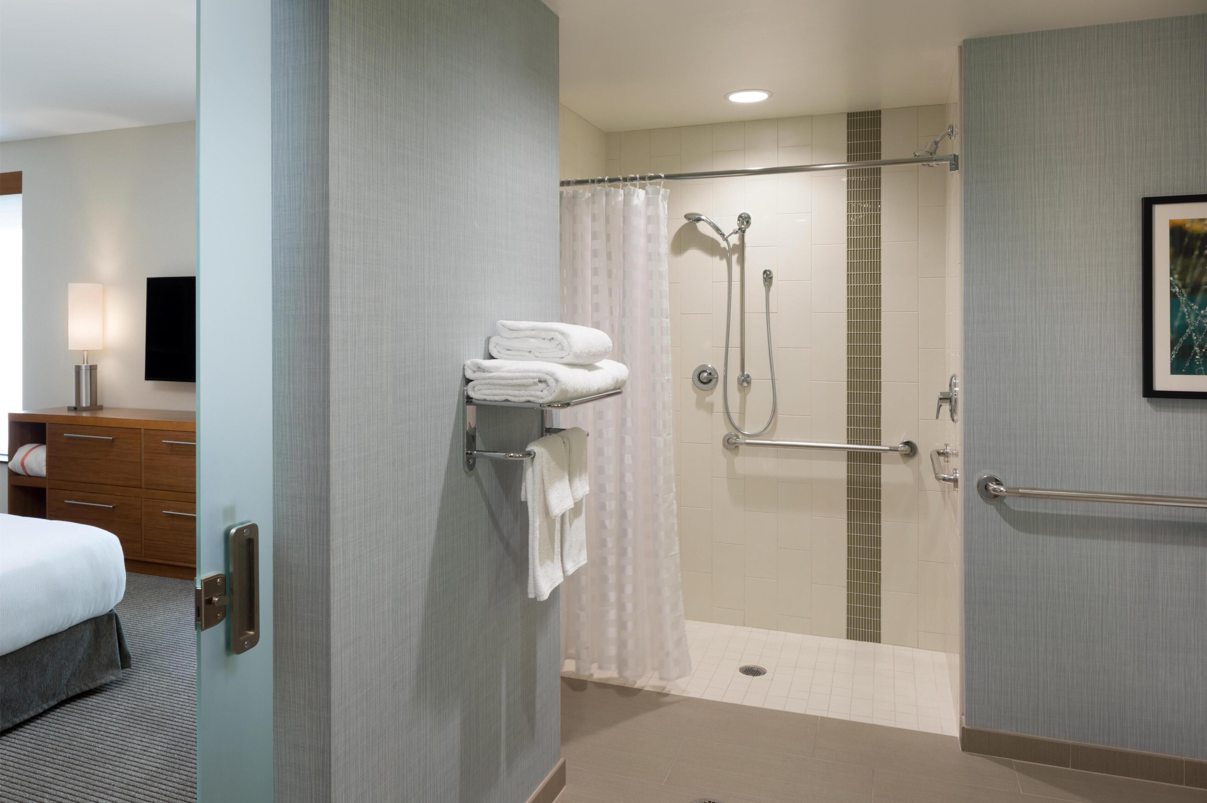 A hotel bathroom with a shower, a towel rack, and a bed.