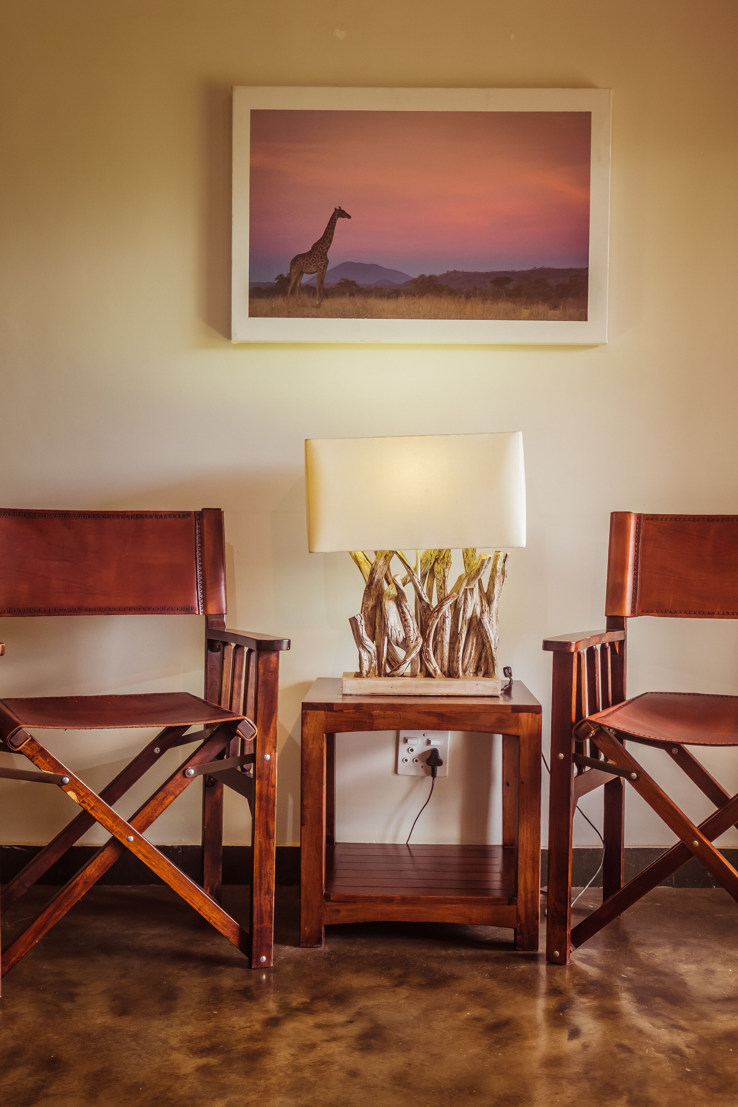 A room with two wooden chairs, a small table with a lamp, and a framed picture of a giraffe on the wall.