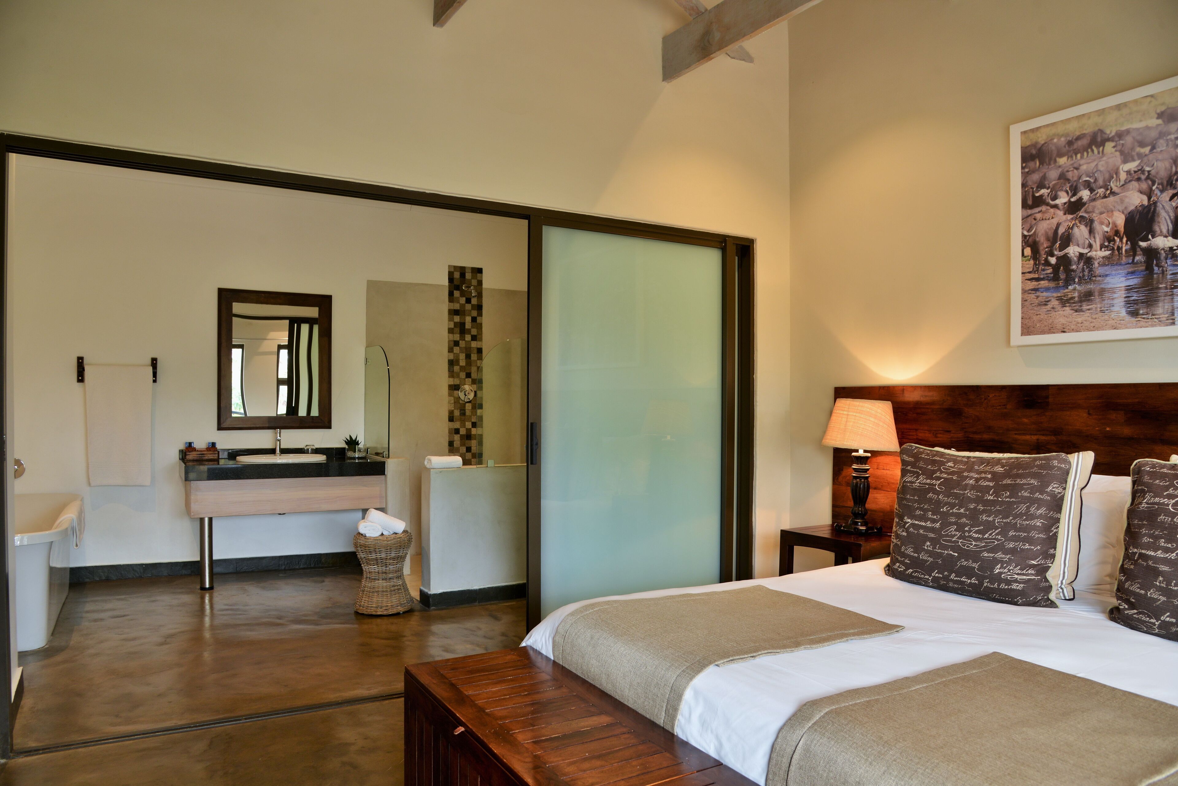 Habitación de hotel con cama, mesita de noche con lámpara, baño con lavabo y espejo, y un cuadro grande en la pared.