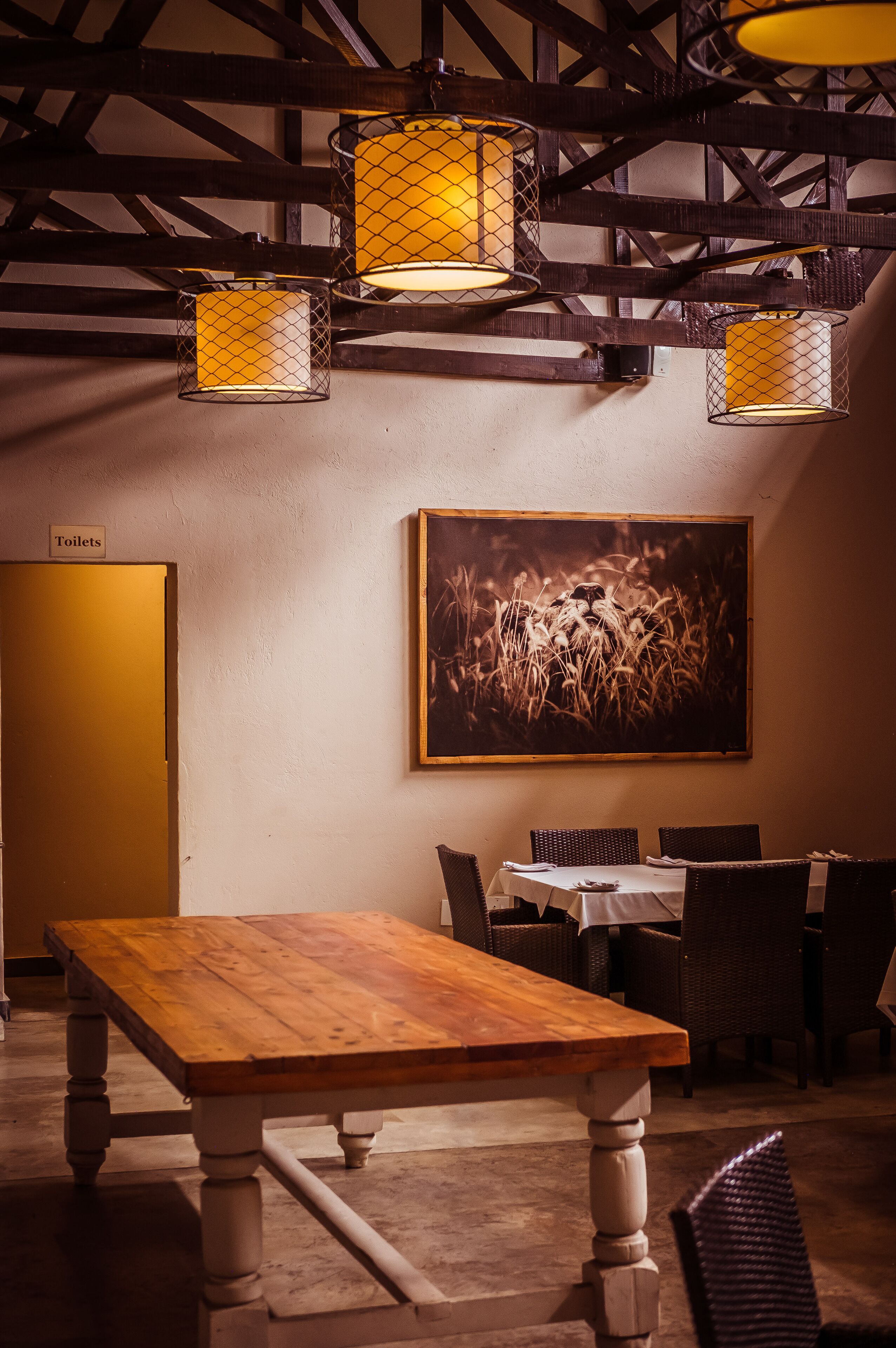 Un comedor con mesa de madera, sillas de mimbre e iluminado por lámparas colgantes estilo farol. Hay un cuadro en la pared y una señalización indicando los servicios.