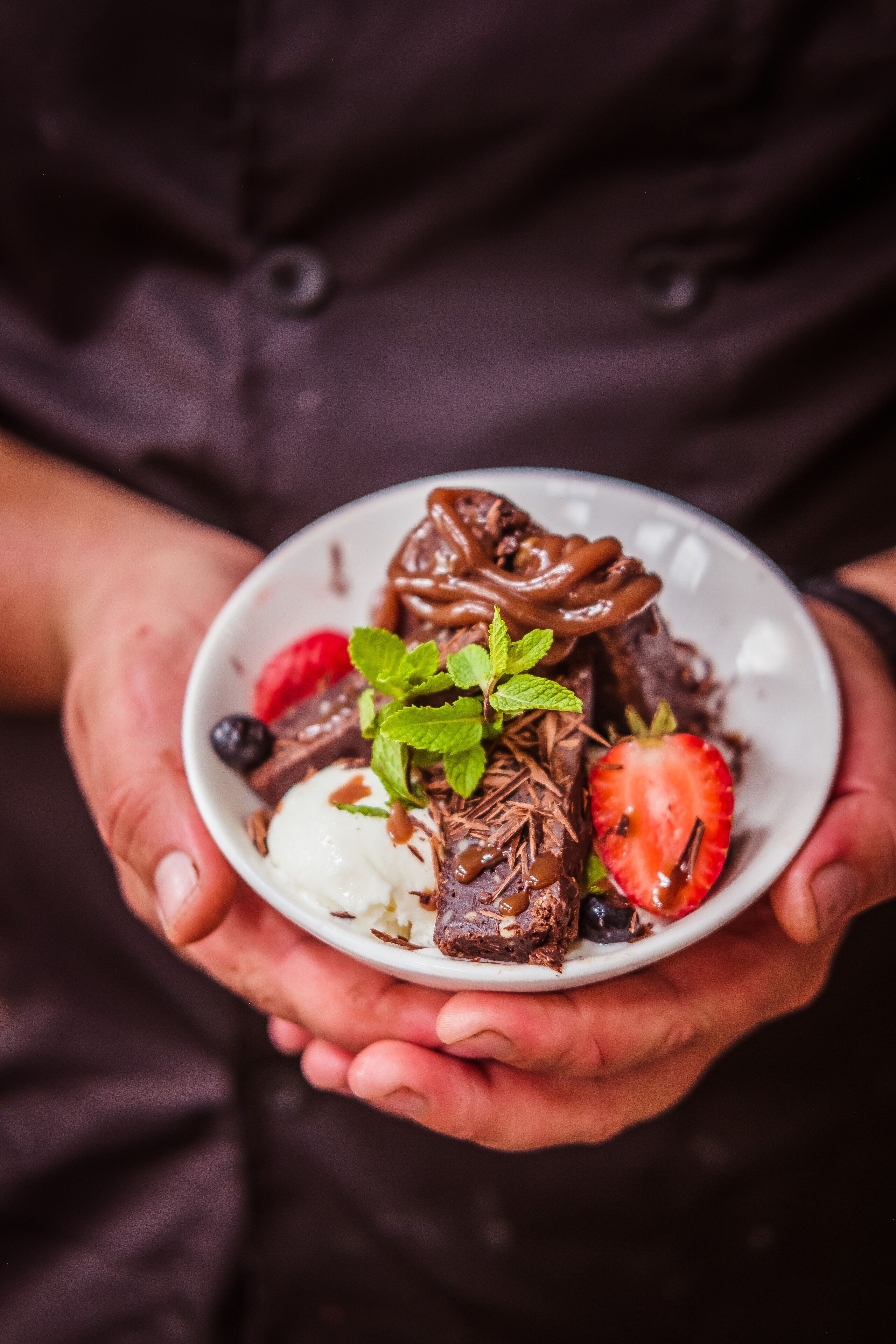 A person holding a bowl of dessert with chocolate, strawberries, and mint.