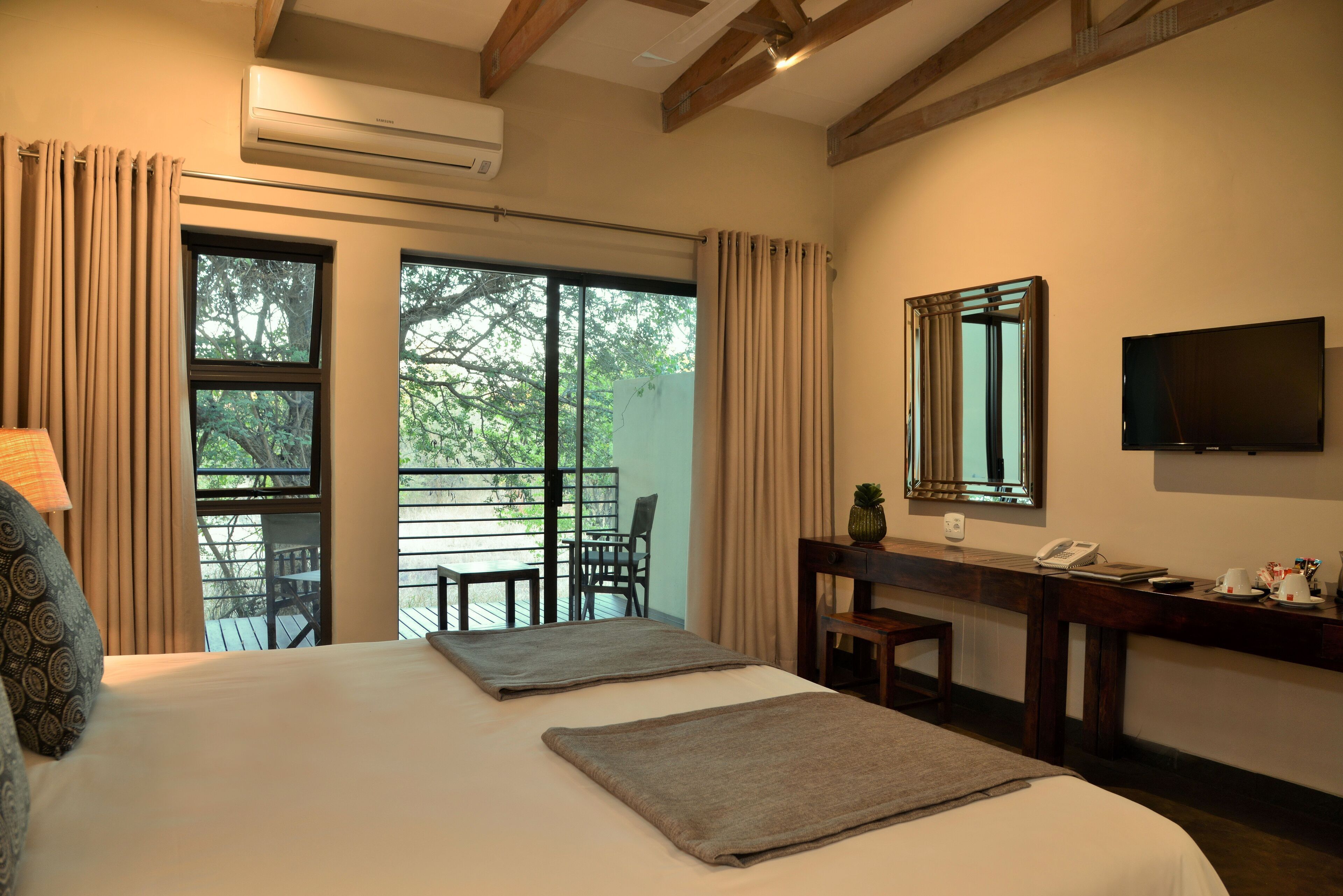 A hotel room with a large bed, a desk with a telephone, a mirror, and a TV. There is a balcony with a chair and a table.