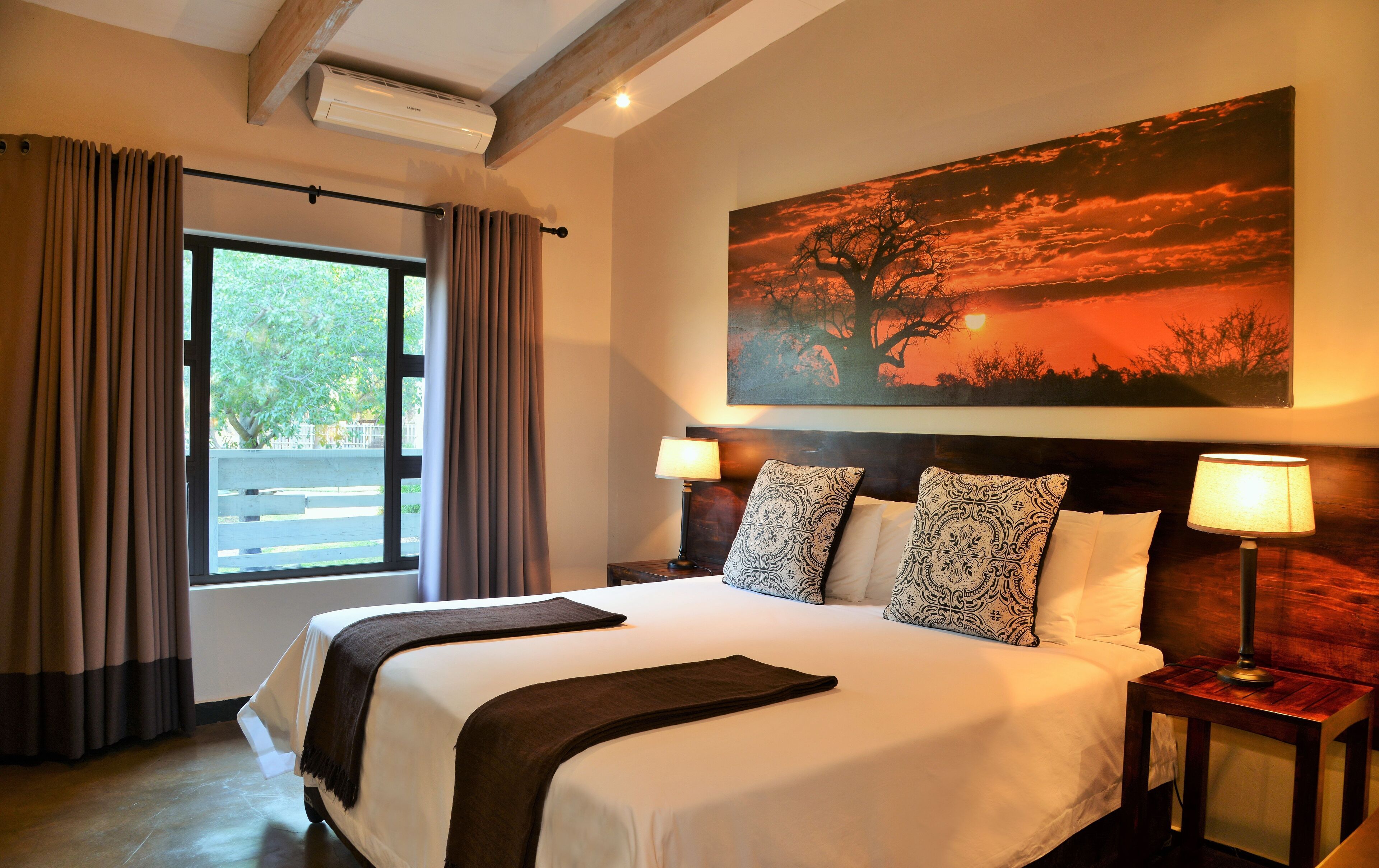 A hotel room with a large bed, two bedside lamps, a painting of a sunset, and a window with curtains.