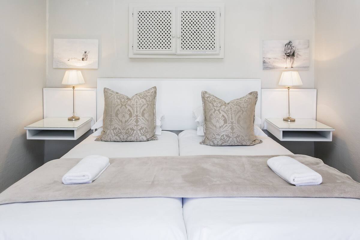 A double bed with two nightstands, each with a lamp and a small table. There are two pillows on the bed, and a blanket is folded at the foot of the bed.