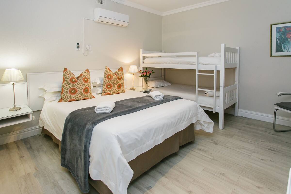 A hotel room with a bunk bed, a bedside table with a lamp, a bedside plant, and a wall-mounted air conditioner.