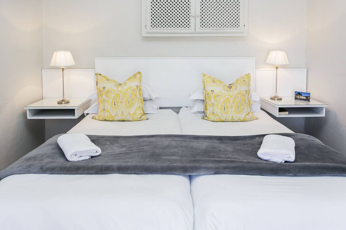 A double bed with two pillows, a grey blanket, and two white towels. There are two bedside tables with lamps and a small card on one.