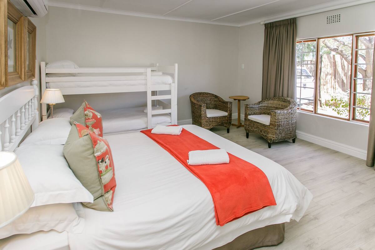 A hotel room with a bunk bed, a single bed, a wicker chair, a small table, and a window with a view of the outside.