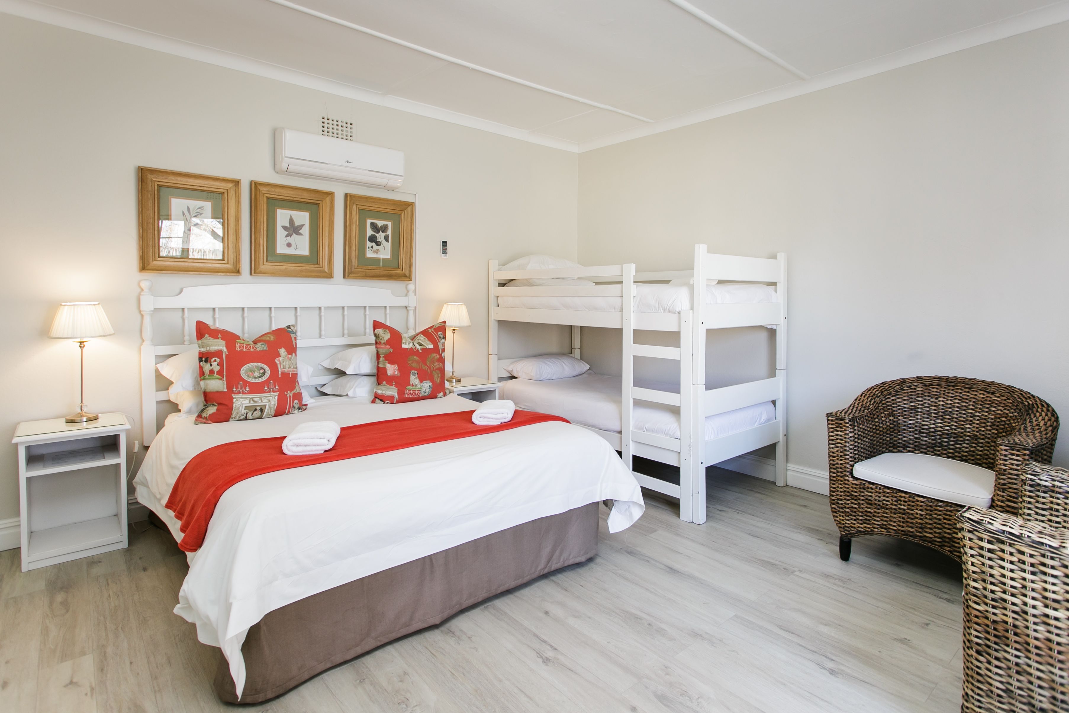 A bedroom with a bunk bed, a single bed with red and white pillows, a wicker chair, and a bedside table with a lamp.