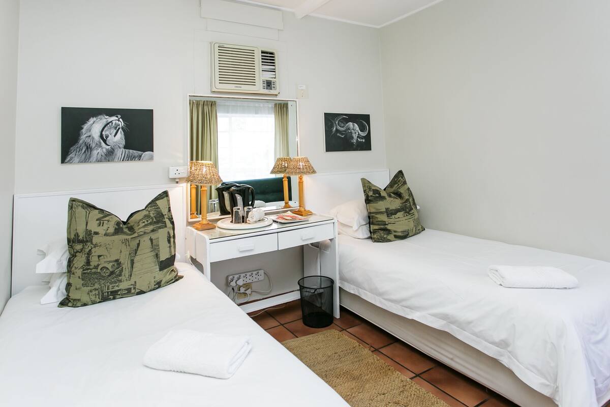 A hotel room with two single beds, a desk with a lamp, a mirror, and two framed pictures on the wall.