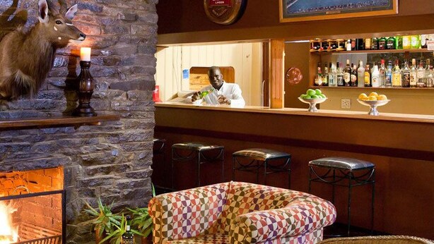 A stone fireplace with a deer head mounted above it, a bar area with stools, and a checkered armchair. - Wagon Wheel Country Lodge (Beaufort West)