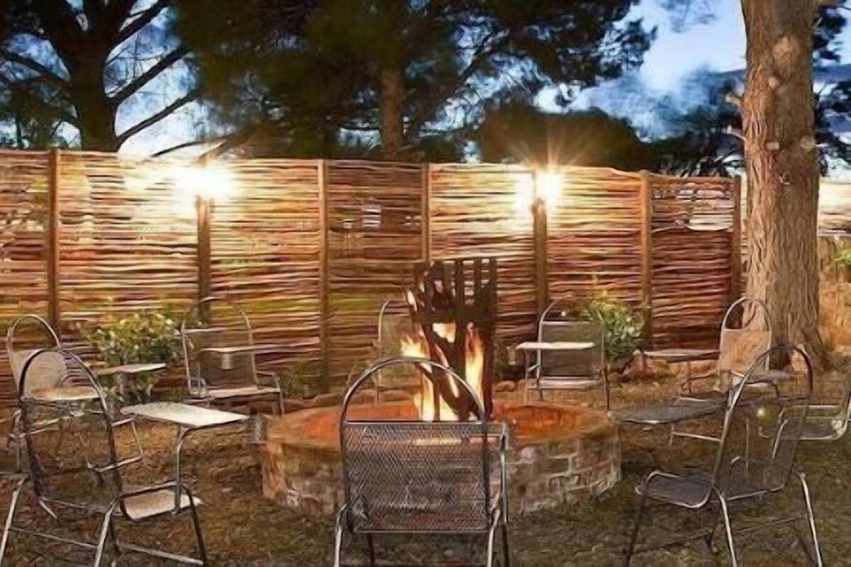 A rustic outdoor seating area with metal chairs and a fire pit, surrounded by wooden fences and a stone structure with a chimney.
