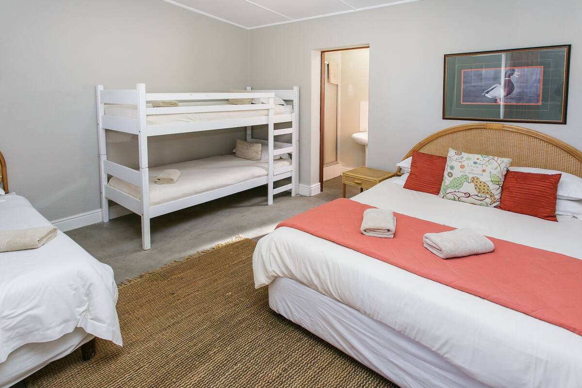 A room with bunk beds, a single bed, a wicker headboard, a framed picture, and a small table with a lamp.